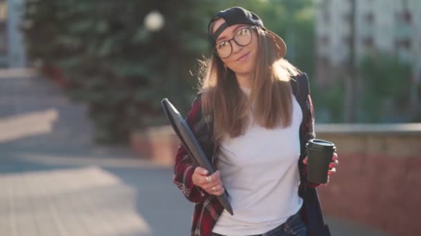 Šťastná mladá studentka oblečená v neformálním oblečení s šálkem kávy a batohem za zády procházející se po městě. Studentka drží notebook a pije kávu. Letní západ slunce. — Stock video