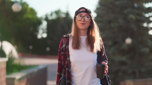 Jovem estudante feliz vestida com roupas casuais com xícara de café e mochila nas costas andando pela cidade. Estudante mulher segurando laptop e beber café. Hora do pôr-do-sol . — Vídeo de Stock