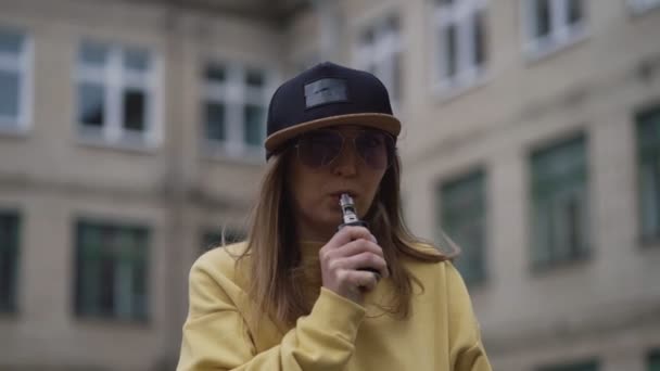 Mooie jonge hipster vrouw in zwarte pet gele hoodie vape ecig. Meisje vape populair ecig gadget, dampen apparaat.Gelukkig brunette vaper meisje met e-cig. Portret van roker vrouwelijk model met elektronische — Stockvideo