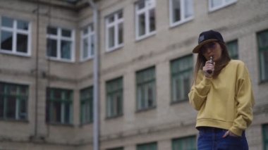 Siyah şapkalı, sarı kapüşonlu, genç ve güzel bir hippi kadın. Kız Vape popüler ecig aleti, Vaping cihazı. E-sigaralı mutlu esmer vampir kız. Sigara içen kadın modelin elektronik portresi