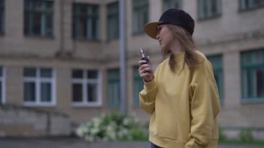 Siyah şapkalı, sarı kapüşonlu, genç ve güzel bir hippi kadın. Kız Vape popüler ecig aleti, Vaping cihazı. E-sigaralı mutlu esmer vampir kız. Sigara içen kadın modelin elektronik portresi