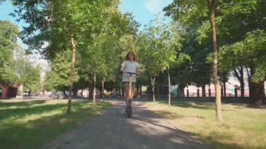 Kız gün batımında şehir parkında elektrikli scooter ya da e-scooter kullanıyor. Şehir parkında elektrikli taşıma kullanan kadın