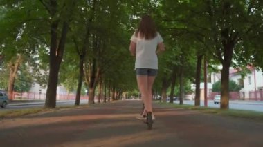 Kız gün batımında şehir parkında elektrikli scooter ya da e-scooter kullanıyor. Şehir parkında elektrikli taşıma kullanan bir kadın. Ön görünüm.