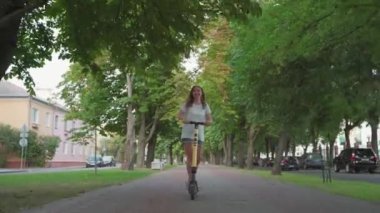 Kız gün batımında şehir parkında elektrikli scooter ya da e-scooter kullanıyor. Şehir parkında elektrikli taşıma kullanan bir kadın. Ön görünüm.