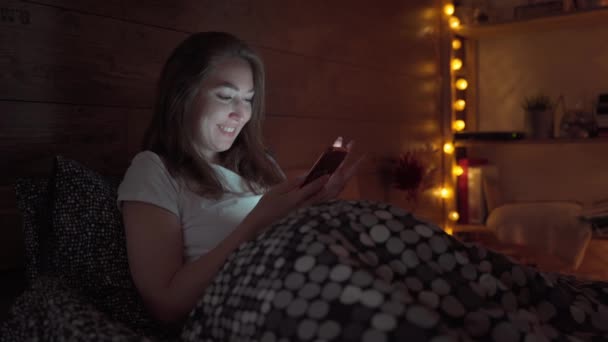 幸せな笑顔の若い女性は夜に自宅のベッドルームでベッドの上でスマートフォンにテキストメッセージ。テクノロジー、インターネット、コミュニケーション、人々の概念は . — ストック動画