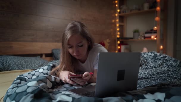 かなり若い女性は、寝室のベッドの上にスマートフォンやラップトップで時間を費やしています。家のコンセプトからの仕事. — ストック動画
