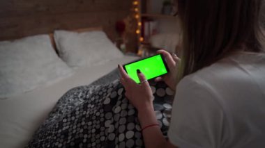 Genç bir kadın cep telefonuyla yatağında dinleniyor. Yatay yeşil akıllı telefon ekranı kullanan bir kız modeli. Kadın cep telefonunun dokunmatik ekranına tıklıyor.