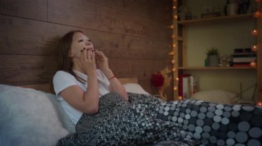 Güzel genç bir kadın sabah yatağında oturuyor ve yüz güzelliği kozmetik nemlendirici maske takıyor. Cilt bakımı kavramı.