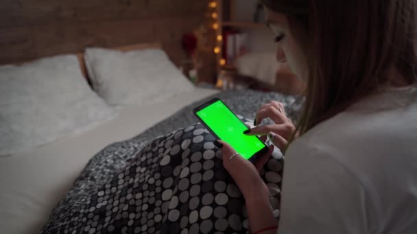Mujer joven relajándose en su cama con su teléfono móvil. Burla de chica usando la pantalla verde del teléfono inteligente. Hembra clics dedo en la pantalla táctil del teléfono celular. — Vídeos de Stock