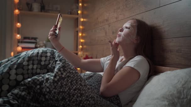 얼굴을 적신 아름다운 화장품 마스크 시트를 착용하고 있는 아름다운 젊은 여성은 저녁 집의 부드러운 침대에서 스마트폰을 사용 한다. 피부 관리 개념. — 비디오