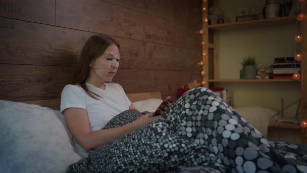 Vacker ung kvinna med ansiktsvatten återfuktande skönhet kosmetisk mask blad använda smartphone i mjuk säng på kvällen hemma. Hudvårds koncept. — Stockvideo