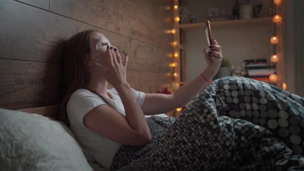 Vacker ung kvinna med ansiktsskönhet fuktgivande kosmetisk mask blad använda smartphone i mjuk säng på kvällen hemma. Hudvårds koncept. — Stockvideo