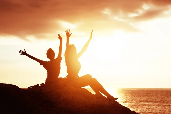 Twee gelukkige meisjes met opgeheven handen tegen zonsondergang zee — Stockfoto