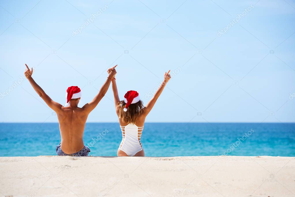 Happy couple sits white beach in christmas hats