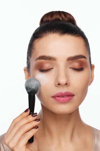 Портрет красивой молодой женщины, держащей кисть в руке и наносящей порошок на лицо кистью. Белый фон
