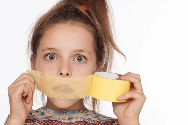 Retrato de uma menina emocional de 8 anos, com o cabelo levantado e em uma blusa com ornamentos que colam fita de papel na boca — Fotografia de Stock