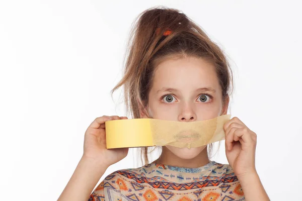 Retrato de uma menina emocional de 8 anos, com o cabelo levantado e em uma blusa com ornamentos que colam fita de papel na boca — Fotografia de Stock