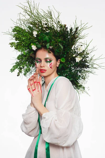 Retrato de una hermosa mujer en un vestido blanco con una cinta verde y con ramas en la cabeza. Maquillaje creativo — Foto de Stock