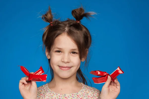 Retrato de uma menina bonita de cinco anos com um penteado interessante e com doces em suas mãos em um fundo azul — Fotografia de Stock