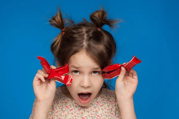Retrato de uma menina bonita de cinco anos com um penteado interessante e com doces em suas mãos em um fundo azul — Fotografia de Stock
