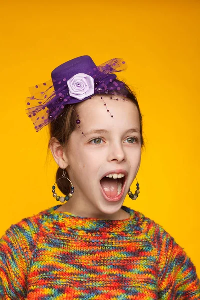 Retrato de uma menina alegre de 8 anos em um chapéu em miniatura e em uma camisola multicolorida. Fundo amarelo — Fotografia de Stock