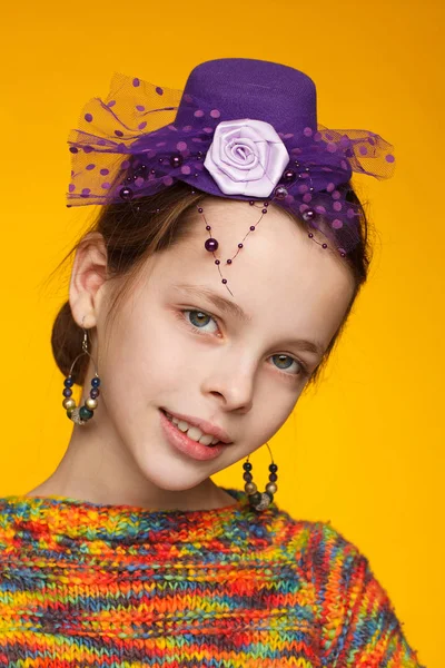 Retrato de uma menina alegre de 8 anos em um chapéu em miniatura e em uma camisola multicolorida. Fundo amarelo — Fotografia de Stock