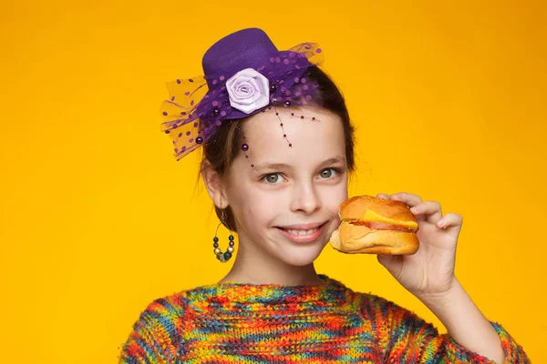 Retrato de uma menina alegre de 8 anos em um chapéu em miniatura, uma camisola multicolorida e com um sanduíche na mão — Fotografia de Stock