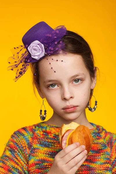 Minyatür şapkalı, çok renkli bir kazak ve elinde sandviç olan 8 yaşındaki neşeli bir kızın portresi