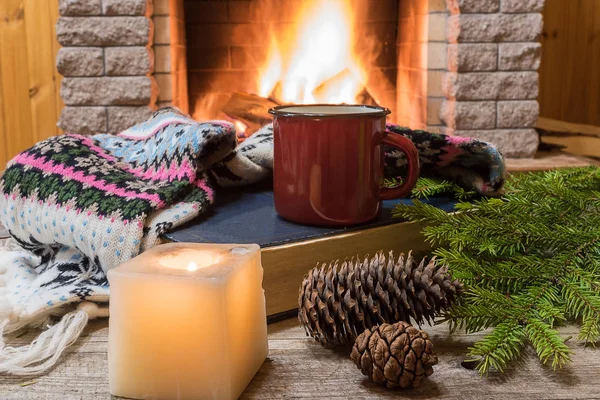 Red enameled mug with tea, and warm wool scarf , a book , near cozy fireplace, in country house, winter vacation.