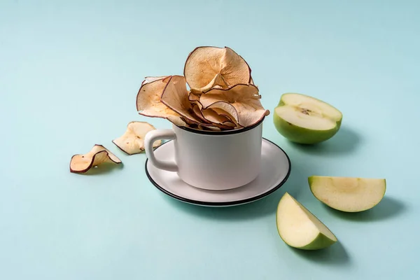 Dried apple chips in a cup and fresh apples over light blue background.