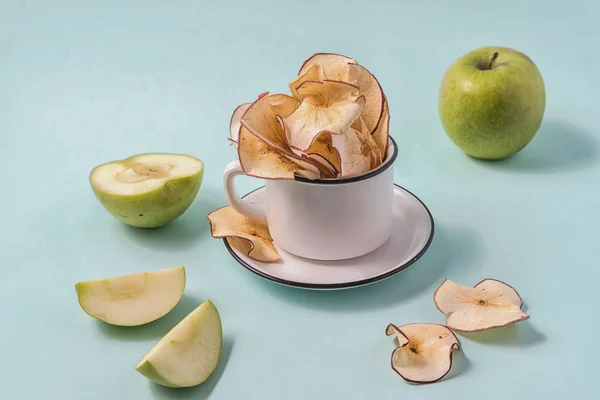 Dried apple chips in a cup and fresh apples over light blue background.