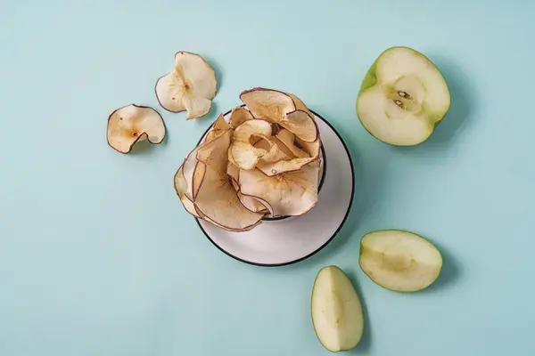 Dried apple chips in a cup and fresh apples on light blue background.