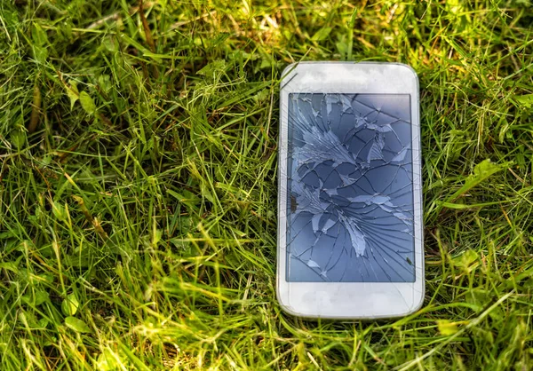 Broken mobile phone with cracked display on green grass background, outdoor, natural light.
