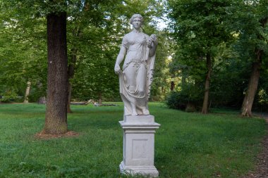 Taş bir kaideye monte edilmiş antik beyaz mermer bir heykelin dikey çekimi. Yeşil, gölgeli tarihi bir parkta geçiyor. Heykel muhtemelen bir periyi, tanrıçayı ya da klasik tarzda alegorik bir figürü tasvir ediyor.