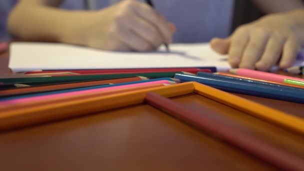 Цветные карандаши разбросаны по столу. Из фокуса детской руки, которая рисует в альбоме — стоковое видео