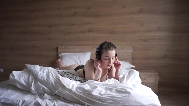 Молодая женщина в нижнем белье слушает музыку с наушниками, лежащими на кровати — стоковое видео