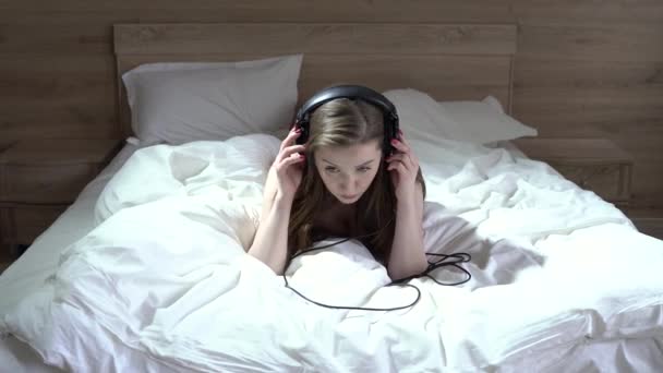 Сексуальная девушка в нижнем белье лежит на кровати и слушает музыку в больших наушниках — стоковое видео