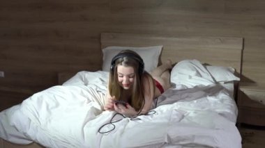 Iç çamaşırı seksi kız, telefon kullanarak ve büyük kulaklıklar müzik dinlerken, yatakta yatıyor