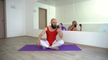 Genç bir neşeli adam bir lastik fitness Mat bir Lotus pozisyonda oturan dumbbells ile ellerinin kasları sallar.