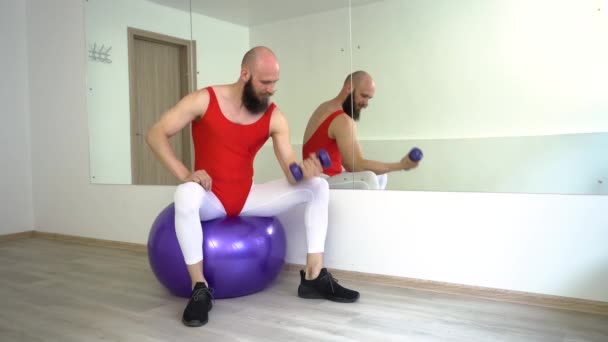 A man in sports leggings pumps the muscles of his hands, sitting on a sports rubber ball. — Stock Video