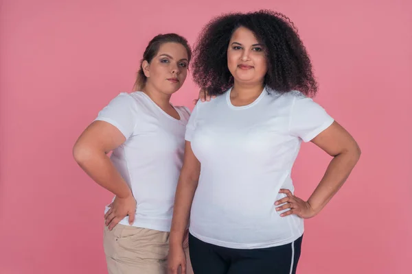 Portrait of multinational girl and Caucasian girl in white t-shirts on pink background. Women with large weight