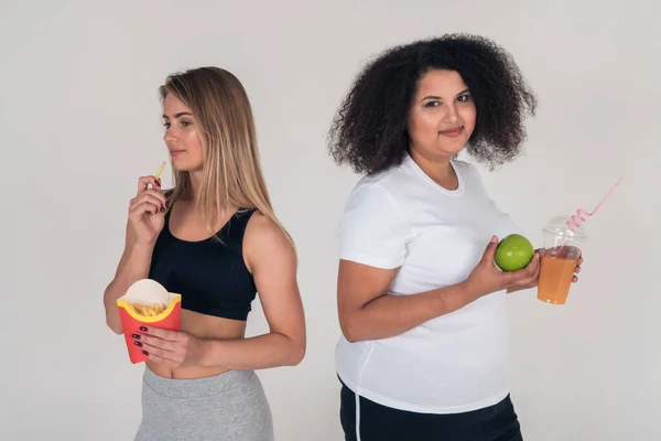 痩せたブロンドがフランスのフライドポテトとぽっちゃりアフリカ系アメリカ人の女の子を食べ、アップルを持っていると白の背景に新鮮な絞り汁の肖像画。体重減少と体重増加 — ストック写真