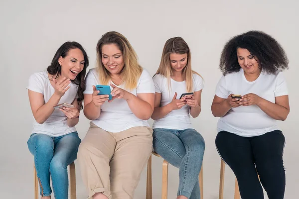 Two girls watch a video on the phone and laugh while two girls look into their phones sitting on a white background