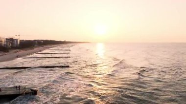 Koobrzeg üzerinde günbatımı antenleri: parlayan gökyüzü ve deniz, uzakta deniz feneri, kumlu sahil altın saatin tadını çıkaran insanlarla dolu.