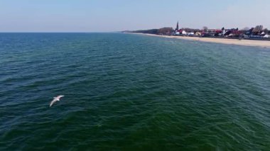 Sarbinowo 'daki Baltık kıyı şeridi Mart ayı güneşi altında parıldıyor: altın plaj, sakin dalgalar, gezinti güvertesi ve köyün sahil evleri arasında yükselen kırmızı tuğlalı kilise.