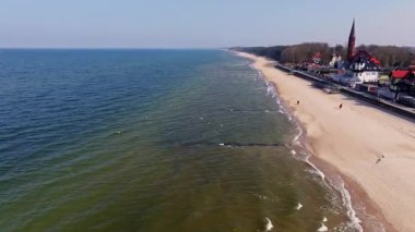 Sarbinowo 'nun kıyı şeridi Mart ayında parlıyor: geniş kumlu sahil, sakin deniz, gezinti güvertesi ve köy merkezindeki zamansız kırmızı tuğlalı kilise..