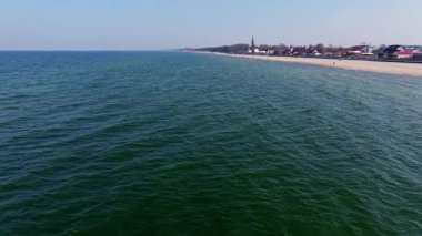 Sarbinowo on a calm spring day: wide sandy beach, golden light, promenade by the sea, and a historic red-brick church at the heart of the village.