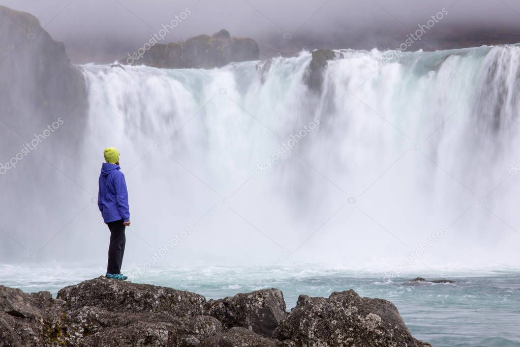 A beautiful young woman is standing against the backdrop of a beautiful waterfall. Iceland
