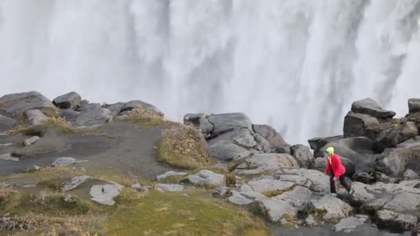 若い女性は 岩のエッジに沿って大きく落ちる強力な荒れ狂う滝を賞賛します 氷河の水の透き通った流れ落ちる崖の上 — ストック動画