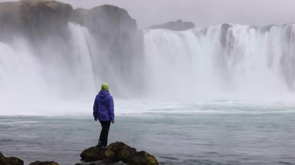 一个年轻的女人钦佩一个强大的狂暴瀑布 沿着岩石边缘严重下降 清澈的冰川水流落在悬崖上 — 图库视频影像